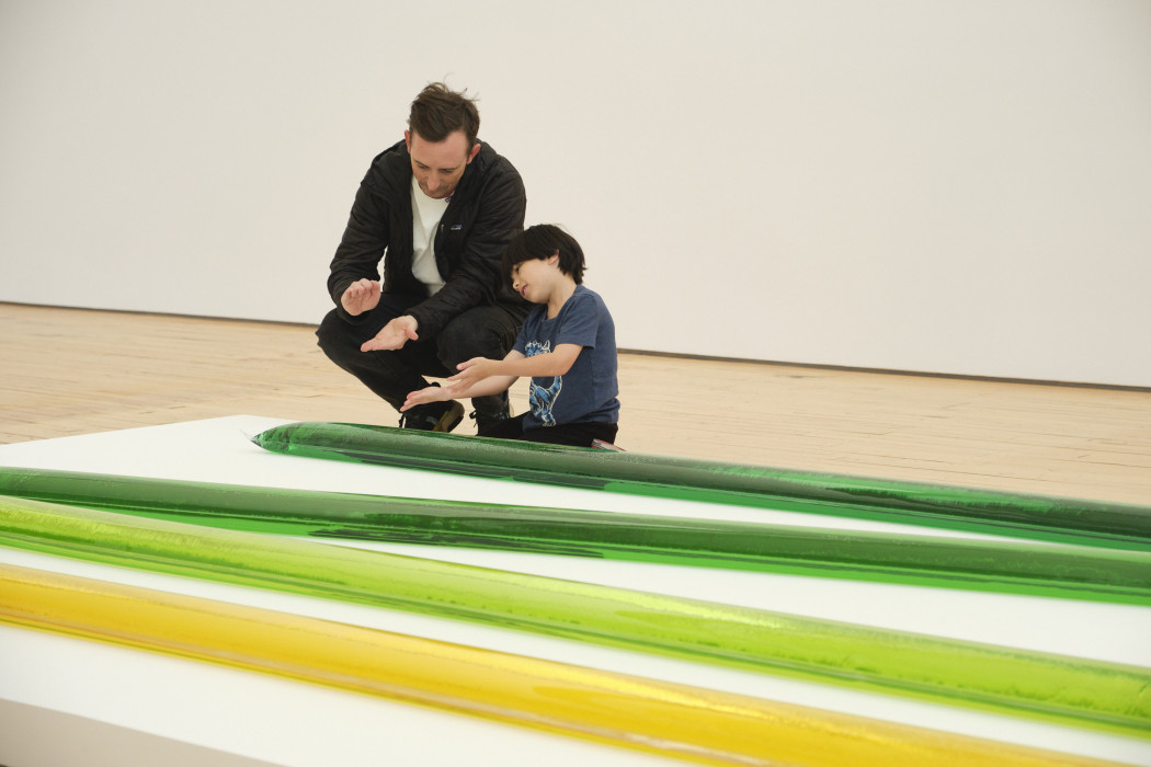 The sculpture is made from plastic and colored water in four different colors, starting closest to the camera is a light yellow, a light yellow-green, a light green, and a dark green. A father and son kneel down to be near a sculpture which lays of a slightly raised platform.
